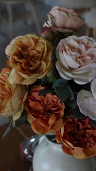 A bouquet of artificial flowers consisting of roses in various shades of orange, peach, and white is arranged in a white ceramic vase. The background is blurred, adding a sense of depth and focus to the flowers.