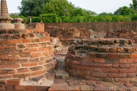 Ancient brick structures with circular bases appear to be partially restored or preserved ruins. Lush green trees and foliage surround the site, suggesting a historic site set within a natural setting. The weathered appearance of the bricks indicates significant age.