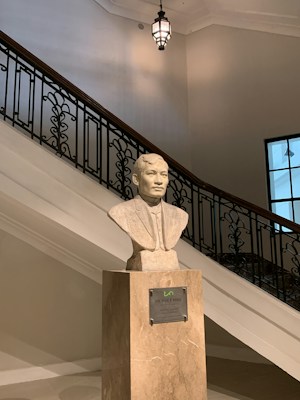 A bust of a person is placed on a pedestal with a plaque. The background features a staircase with ornate railings and a hanging light fixture above. The setting appears to be indoors, with ambient lighting and a window visible on the right side.