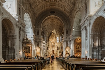 A grand, ornate church interior featuring an arched ceiling with intricate designs and large columns. Elaborate wooden pews line the nave, leading towards the altar adorned with religious artwork and sculptures. Soft lighting enhances the architectural details and the subdued atmosphere. People are scattered throughout, some seated and others walking towards the altar.