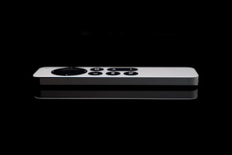 A sleek white remote control with a circular directional pad and several buttons is positioned against a dark, reflective surface. The minimalistic design is accentuated by the sharp contrast between the white remote and the black background.
