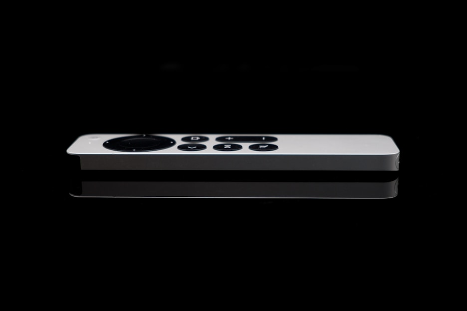 A sleek white remote control with a circular directional pad and several buttons is positioned against a dark, reflective surface. The minimalistic design is accentuated by the sharp contrast between the white remote and the black background.