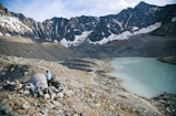 A small campsite is set up on rocky terrain near a turquoise mountain lake, surrounded by towering snow-capped peaks. A person in outdoor gear stands near a tent, looking towards the lake. The sky is partly cloudy, casting a serene atmosphere over the scene.