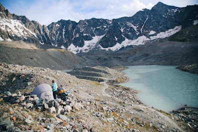 A small campsite is set up on rocky terrain near a turquoise mountain lake, surrounded by towering snow-capped peaks. A person in outdoor gear stands near a tent, looking towards the lake. The sky is partly cloudy, casting a serene atmosphere over the scene.