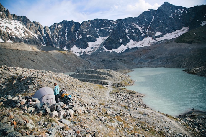 A small campsite is set up on rocky terrain near a turquoise mountain lake, surrounded by towering snow-capped peaks. A person in outdoor gear stands near a tent, looking towards the lake. The sky is partly cloudy, casting a serene atmosphere over the scene.