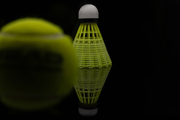 A close-up view of a neon yellow shuttlecock with a black background. The shuttlecock is positioned upright, with its reflection visible on the surface below. To the side, there is a blurred view of a neon yellow tennis ball, partially obstructed and out of focus.