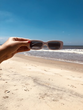 Hands holding a pair of sleek sunglasses with a blurred beach background.
