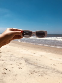 A hand holding a pair of white-framed sunglasses is positioned in front of a beach scene, with sandy shores and gentle waves in the background under a clear blue sky.