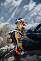 A pair of yellow and black mountaineering boots with crampons attached, resting on rocky terrain. The background shows a blurred, snowy mountain landscape, indicating a high-altitude environment.