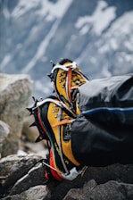 A pair of yellow and black mountaineering boots with crampons attached, resting on rocky terrain. The background shows a blurred, snowy mountain landscape, indicating a high-altitude environment.