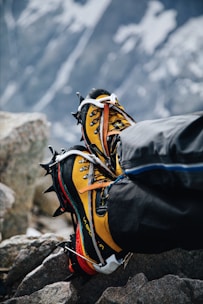 A pair of yellow and black mountaineering boots with crampons attached, resting on rocky terrain. The background shows a blurred, snowy mountain landscape, indicating a high-altitude environment.