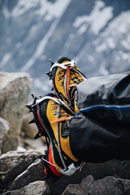 A pair of yellow and black mountaineering boots with crampons attached, resting on rocky terrain. The background shows a blurred, snowy mountain landscape, indicating a high-altitude environment.