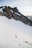 A rugged mountain landscape with steep, jagged peaks covered in patches of snow. Two climbers with gear are ascending a snow-covered slope, surrounded by impressive rock formations under a pale sky.