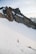 A rugged mountain landscape with steep, jagged peaks covered in patches of snow. Two climbers with gear are ascending a snow-covered slope, surrounded by impressive rock formations under a pale sky.