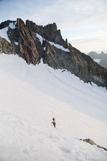 A rugged mountain landscape with steep, jagged peaks covered in patches of snow. Two climbers with gear are ascending a snow-covered slope, surrounded by impressive rock formations under a pale sky.