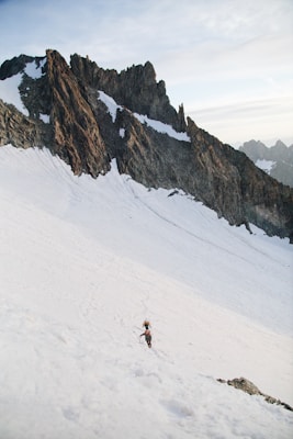 A rugged mountain landscape with steep, jagged peaks covered in patches of snow. Two climbers with gear are ascending a snow-covered slope, surrounded by impressive rock formations under a pale sky.
