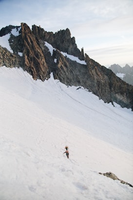 A rugged mountain landscape with steep, jagged peaks covered in patches of snow. Two climbers with gear are ascending a snow-covered slope, surrounded by impressive rock formations under a pale sky.