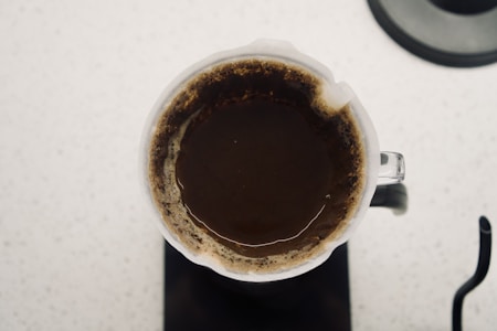 A top-down view of a coffee brewing process using a glass pour-over coffee maker. The coffee grounds are saturated, forming a dark brown color with lighter edges visible around the perimeter. The coffee maker sits on a white speckled surface.