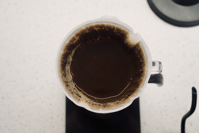 A top-down view of a coffee brewing process using a glass pour-over coffee maker. The coffee grounds are saturated, forming a dark brown color with lighter edges visible around the perimeter. The coffee maker sits on a white speckled surface.