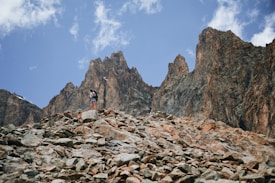 A rugged mountain landscape with a hiker standing on a rocky terrain. The hiker is wearing a backpack and appears to be taking in the majestic scenery. Jagged peaks rise sharply in the background under a blue sky with scattered clouds.