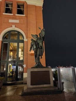 A statue depicting an angelic figure with wings embracing a person stands prominently on a stone pedestal in front of a red brick building with large windows. The scene is set against a night sky with city lights visible in the distance.