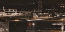 A nighttime view of an airport with multiple airplanes parked near the terminal. The airport is illuminated with numerous bright lights, and there are visible control towers and surrounding buildings. Light trails indicate moving vehicles or aircraft, adding a dynamic element to the scene.
