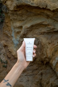 Close-up of a hand holding a jar of agave skincare cream with agave plants in the background.