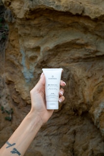 A hand with a tattoo on the wrist is holding a tube of hand cream labeled 'PLANTADEA BIO CREMA MANI' against a textured rocky background.