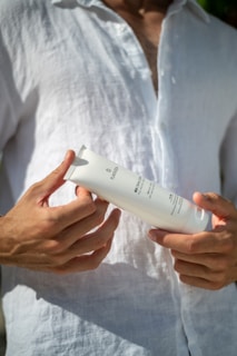 A person is holding a white tube of skincare or cosmetic product, with their fingers lightly gripping the item. They are wearing a white button-down shirt, and their hands are prominently featured against the fabric.