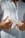 A person is holding a white tube of skincare or cosmetic product, with their fingers lightly gripping the item. They are wearing a white button-down shirt, and their hands are prominently featured against the fabric.