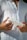 A person is holding a white tube of skincare or cosmetic product, with their fingers lightly gripping the item. They are wearing a white button-down shirt, and their hands are prominently featured against the fabric.