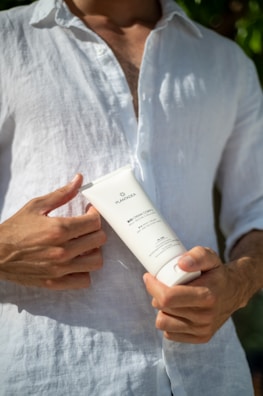 A person wearing a white linen shirt is holding a white tube of bio body cream closer to their chest. The person is seen from the neck to the waist, and there is some green foliage in the blurred background.