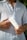 A person wearing a white linen shirt is holding a white tube of bio body cream closer to their chest. The person is seen from the neck to the waist, and there is some green foliage in the blurred background.