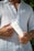 A person wearing a white linen shirt is holding a white tube of bio body cream closer to their chest. The person is seen from the neck to the waist, and there is some green foliage in the blurred background.