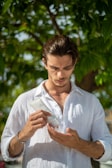 A person stands outdoors under a tree, holding a tube of skincare product and inspecting it closely. Wearing a white shirt, the individual is focused on applying or examining the product in their hand, set against a backdrop of green leaves.