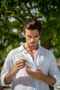 A person stands outdoors under a tree, holding a tube of skincare product and inspecting it closely. Wearing a white shirt, the individual is focused on applying or examining the product in their hand, set against a backdrop of green leaves.