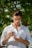 A person stands outdoors under a tree, holding a tube of skincare product and inspecting it closely. Wearing a white shirt, the individual is focused on applying or examining the product in their hand, set against a backdrop of green leaves.