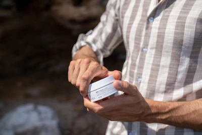 Close-up of a hand holding a Safira cream jar with natural ingredients softly blurred in the background.