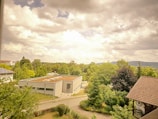 A scenic view featuring a mix of residential and natural landscapes. A modern flat-roofed building and traditional house with a tiled roof sit amidst lush green trees under a sky filled with fluffy clouds. The distant hills provide a backdrop to this serene suburban setting.