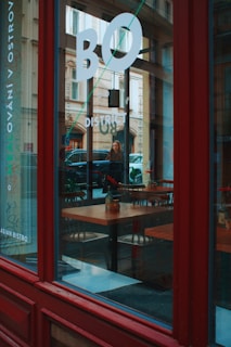 Through a large glass window with bold white lettering, the interior of a bistro is visible, featuring neatly arranged wooden tables and chairs. A vase with a single red flower is placed on a table, adding a touch of color. The reflection on the glass shows a street scene with a parked car and a woman walking by. The exterior is characterized by a rich red frame.