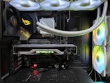 Inside view of a computer case showcasing a sophisticated gaming setup. Visible components include a GeForce RTX graphics card illuminated with green lighting, an ASUS motherboard, cooling fans with LED lighting, and a liquid cooling system with white tubes. Cables are neatly organized, and overall, the hardware suggests a high-performance machine.