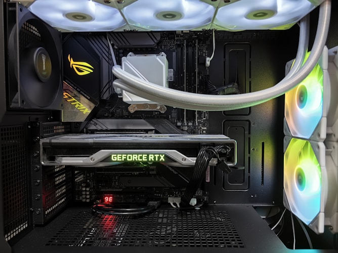 Inside view of a computer case showcasing a sophisticated gaming setup. Visible components include a GeForce RTX graphics card illuminated with green lighting, an ASUS motherboard, cooling fans with LED lighting, and a liquid cooling system with white tubes. Cables are neatly organized, and overall, the hardware suggests a high-performance machine.