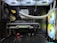 Inside view of a computer case showcasing a sophisticated gaming setup. Visible components include a GeForce RTX graphics card illuminated with green lighting, an ASUS motherboard, cooling fans with LED lighting, and a liquid cooling system with white tubes. Cables are neatly organized, and overall, the hardware suggests a high-performance machine.