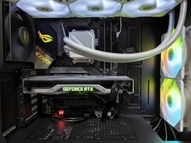 Inside view of a computer case showcasing a sophisticated gaming setup. Visible components include a GeForce RTX graphics card illuminated with green lighting, an ASUS motherboard, cooling fans with LED lighting, and a liquid cooling system with white tubes. Cables are neatly organized, and overall, the hardware suggests a high-performance machine.