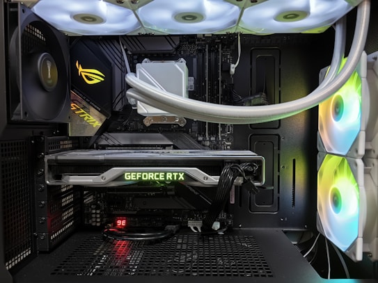 Inside view of a computer case showcasing a sophisticated gaming setup. Visible components include a GeForce RTX graphics card illuminated with green lighting, an ASUS motherboard, cooling fans with LED lighting, and a liquid cooling system with white tubes. Cables are neatly organized, and overall, the hardware suggests a high-performance machine.