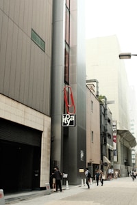 A narrow urban street flanked by modern buildings, featuring a large red paperclip attached to a structure with the word 'lofty' on a sign. Several pedestrians are walking along the sidewalk, and there's a sense of urban hustle. The overall architecture is contemporary with glass windows and various signs. Cones and no parking signs are placed along the edge of the building.