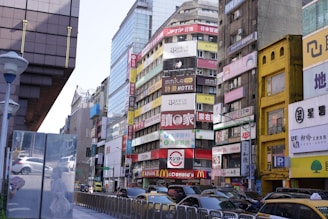 A bustling urban street scene featuring tall buildings with numerous colorful advertisements and signs in various languages. The street is lined with cars, and a McDonald's is prominently visible among the signs. The buildings are modern and densely packed, typical of a busy city environment.