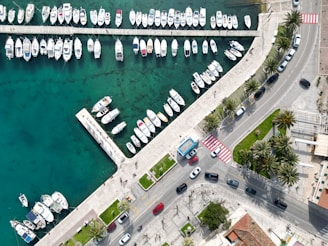 An aerial view of a marina featuring numerous small boats moored along a dock. The water is a deep turquoise, creating a striking contrast with the white boats. Adjacent to the marina is a road lined with palm trees and parked cars. The area includes pedestrian crossings and sidewalks, indicating a waterfront promenade. Green spaces and sidewalks are visible, suggesting a developed urban environment.