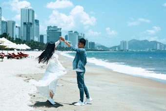 A couple dances joyfully on a sandy beach with a vibrant city skyline in the background. The woman wears a flowing white dress, and the man is dressed in denim. Nearby, red beach chairs and umbrellas line the shore. The ocean's turquoise waves gently lap the sand under a clear blue sky, dotted with a few clouds.