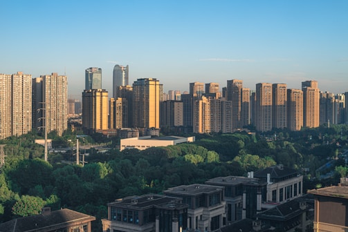 A cityscape featuring a line of tall modern skyscrapers with glass and concrete facades, casting long shadows. Several high-rise apartment buildings cluster together against a clear blue sky. In the foreground, there are lush green trees and a few lower buildings.